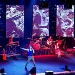 Hudobný koncert kapely s obrazmi vojakov na veľkých obrazovkách.