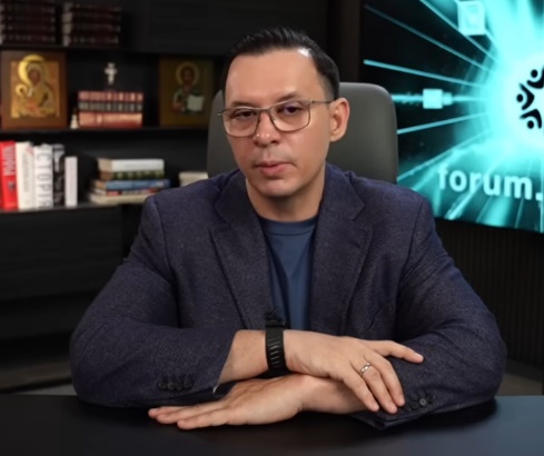Muž v okuliariach pri stole s knihami v pozadí