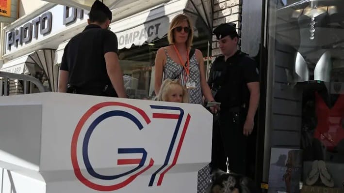 Biely pult G7 s logom, žena s dieťaťom a policajti.